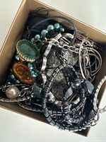 Box filled with assorted jewelry including rings, bracelets, and necklaces showing various materials, colors, and designs.