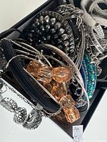 Close-up of assorted silver-tone chain bracelets, metal bangles, faceted amber-colored bead bracelet, black rhinestone circular earrings, and silver-colored pineapple-shaped ring inside a jewelry box.