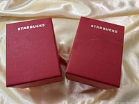 Two red Starbucks boxes containing mugs, with white Starbucks logo on top, shown on a silky background.