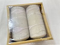 Two white candles in textured cream-colored holders in a cardboard box wrapped in plastic.