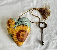 Front of heart shaped decorative lock, painted with sunflowers and 'You Are My Sunshine' text, attached to key with golden tassel.