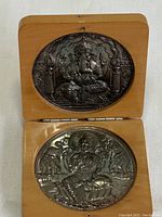 Front inside of wooden hinged display case showing two silver-colored coins with Indian religious motifs.