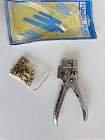 Photo showing punch plier tool and bag of small brass eyelets on white background with original packaging for eyelets visible.