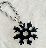 Black snowflake multi-tool attached to a carabiner shown against a white fabric background, highlighting tool shapes and carabiner.