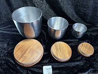 Three metal cups of varying sizes each paired with a round wooden lid, set against a black cloth background.