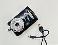 Top and front view of small black digital camera displaying 7.0MP label, lens, and USB cable