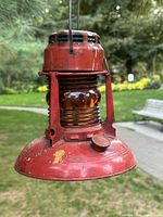 Front and side view of vintage red metal traffic guard lantern with amber ribbed glass globe, set outdoors on a wooden surface with greenery in background.
