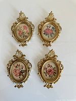Four vintage oval photo frames with gold-tone ornate detailing and floral needlepoint inside, arranged on a white background.