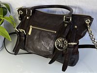 Side view of the brown leather handbag showing the gold zipper pocket and MK pendant logo charm with chain strap.