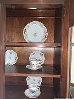 China plates and teacups with floral patterns and gold-colored edges arranged on wooden shelves inside a hutch.