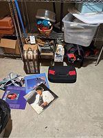 Metal shelving unit with assorted items stored including a vintage map-themed wine trunk, plastic storage containers, a junior metal detector, various kitchen items, and vinyl records spread on the floor.