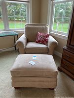Beige La-Z-Boy armchair with rolled arms, cushioned back, red and white patterned decorative pillow, and matching ottoman positioned in front of window.