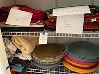Shelves with stacks of folded placemats, table runners, and napkins in various colors and seasonal themes with handwritten labels.