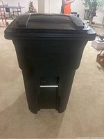 Front view showing the lid, handle and wheel attachment of the black rolling trash can.