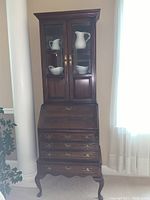 Front view of entire dark wood secretary desk showing glass doors with white ceramic pitchers and bowls inside, drop front desk closed, and four drawers with brass pulls.