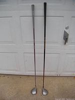 Two Pro Kennex golf drivers standing outside in front of a white door; one has a purple shaft and grey grip (ladies), the other a dark shaft with gold detailing and black grip (mens).