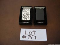 Two Zippo lighters in original packaging side by side on a brown surface. One lighter has a silver filigree pattern, the other is black crackle. Both have 'Made in USA' labels.