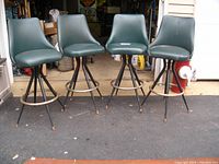Four green vinyl seat bar stools with black metal legs and chrome footrings showing surface rust, sitting on asphalt outside garage.