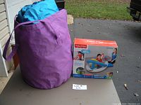Purple fabric bag with handles containing bounce house blower and boxed Fisher-Price Small Ball Pit outside on pavement