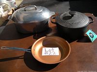 Three cookware items on table: large aluminum roaster with lid, black cast iron Cuisinart Dutch oven, and copper frying pan with black handle.