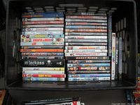 Photo showing DVDs stacked in milk crate with visible titles such as Jackass Number Two, Elf, Cheaper by the Dozen, Ghost Rider, and others.