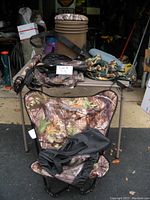 Camouflage folding chair in front of table with bucket, seat cushion, backpack and ground blind bag in background.