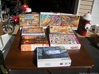 Eight jigsaw puzzles on a table in sunlight. Six are 2000-piece puzzles, two 1000-piece. Visible puzzle images include cats, cabins, shops, and landscapes.