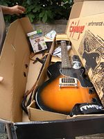 Open box showing the Epiphone Junior electric guitar in sunburst finish, guitar stand, strap, and Rocksmith Xbox game box.