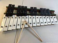 Full Xylophone showing 25 keys alternating black and white metal keys mounted on wooden base with two green support strips. Pair of wooden sticks placed on the keys.
