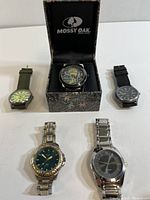 Five assorted wrist watches displayed: Mossy Oak watch in camo box, two on top row with fabric and leather straps, two on bottom row with metal bracelets.