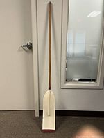 Full length view of the wooden rowing boat oar leaning against a wall. Shows overall length and condition of the shaft and blade.