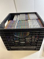 Black plastic milk crate filled with numerous CDs arranged vertically, showing colorful spines.