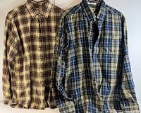 Two men's plaid flannel shirts laid flat, one beige/brown/maroon plaid, one blue/gray/white plaid.