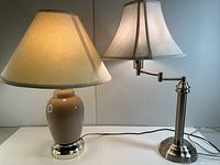 Photo showing both table lamps side by side on a white surface against a neutral background, one with a ceramic base and one with a steel base and adjustable arm.