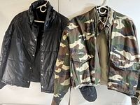 Two women's medium jackets hanging on hangers. One black quilted jacket with snap buttons and one camouflage print jacket with flap pockets and frayed hem.