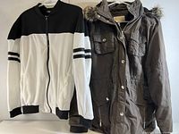Two women's jackets hanging on hangers; left is white and black zip-up jacket, right is brown hooded coat with fur trim and multiple pockets.