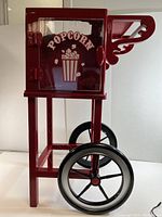 Front and side views of WestBand popcorn cart showing red metal frame, clear door with popcorn graphics, and large black wheels.