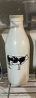 White vintage milk bottle with a black stamped cow image on the front, closed with a screw cap, positioned on a wire rack shelf.