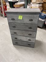 Full view of gray wooden dresser with 4 drawers and metal handles, placed on concrete floor with storage items around.