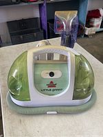 Front view of Bissell Little Green Machine cleaner showing white and green body with hose wrapped around base and purple hand cleaning tool in background.