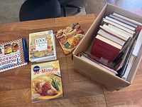 Top view of box with cookbooks inside and several cookbooks placed beside it on a wooden table.