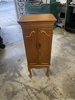 Wood cabinet closed, showing the overall structure with wood finish, decorative legs, and double front doors with carved handles.