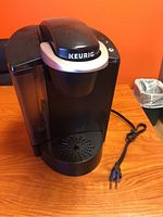 Front-left angle of the black Keurig coffee maker on a wooden table with visible power cord and drip tray.