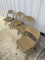 Four beige metal folding chairs arranged side by side on concrete, showing overall set condition and structure.