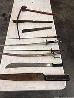 Seven edged weapons laid out on white table: one axe with wooden handle, several swords including one rapier-style, and machetes with signs of rust.