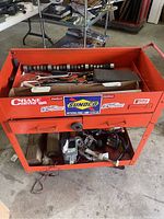 Orange Matco branded mechanics cart with tools and parts on top and bottom shelves.
