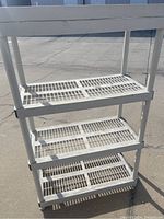 Front view of white plastic storage shelf with four ventilated slatted shelves and tubular supports.