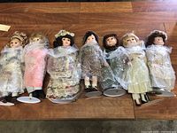 Seven porcelain dolls laid in a row on a wooden surface, each wrapped in bubble wrap and mounted on doll stands.