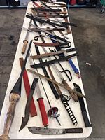 Full table view of assorted swords, machetes, and knives showing various blades and handles, some with sheaths and wrapped grips.