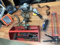 Wide view of assorted tools on a wooden table including small air compressor, electric hand drill, caulking gun, hand crank brace drill, pressure gauges, and a red metal toolbox with various hand tools.